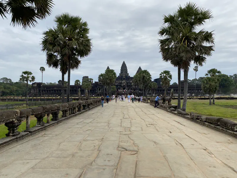 Stone walkway lined with palm trees leading to Angkor Wat temple with tourists approaching.