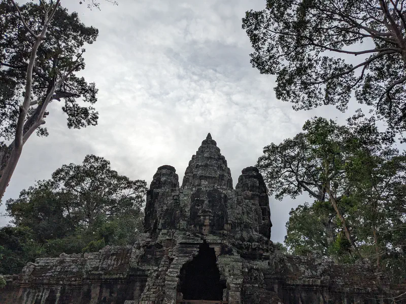 Stone gate with carved faces at Angkor Thom framed by tall trees.