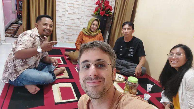 Group of four adults sitting together on a carpeted floor, smiling at the camera during a casual indoor gathering.