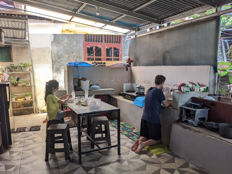 Open-air kitchen with a girl sitting at a table eating and a man washing dishes at the sink, with cooking utensils and pots arranged on the counter.
