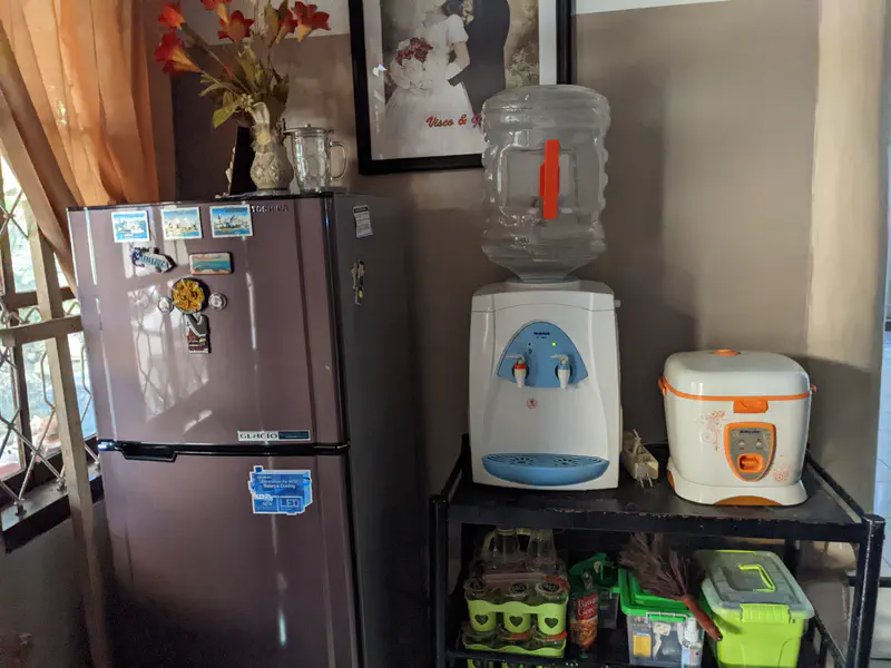 Kitchen corner with a refrigerator decorated with magnets, a water dispenser with a large bottle, and a rice cooker placed on a metal rack storing drinks and supplies.
