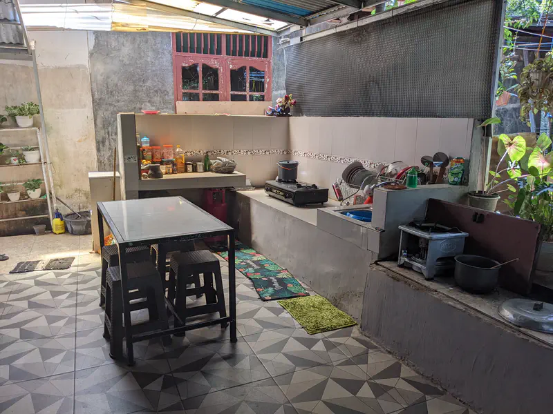 Open-air kitchen with tiled floor, a metal dining table with stools, countertop stove, sink with dishes, and shelves holding cooking supplies.