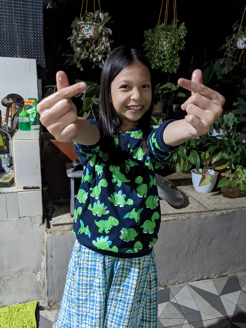 Smiling girl in a dinosaur-print shirt making finger heart gestures with both hands in a garden setting at night.