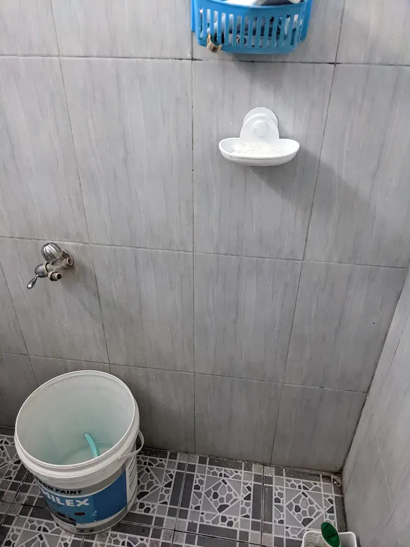 Simple bathroom with tiled walls and floor, showing a bucket with a hose, a wall tap, and a soap dish with a bar of soap.