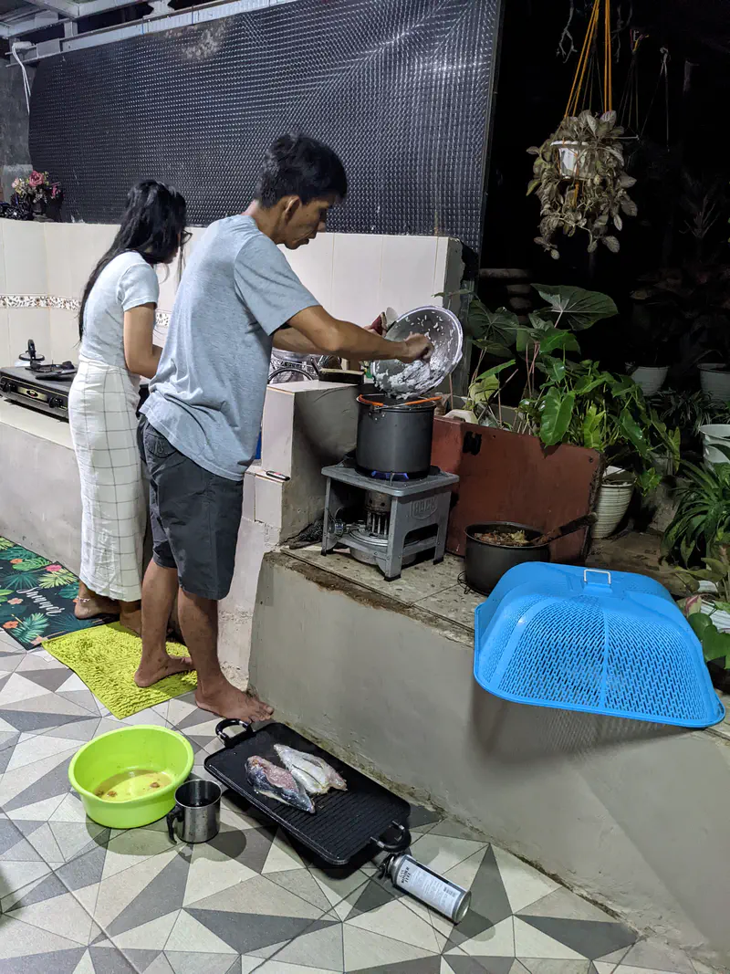 Man cooking over a gas stove while a woman prepares food at the counter in an open-air kitchen, with fish and a bowl of water on the floor nearby.