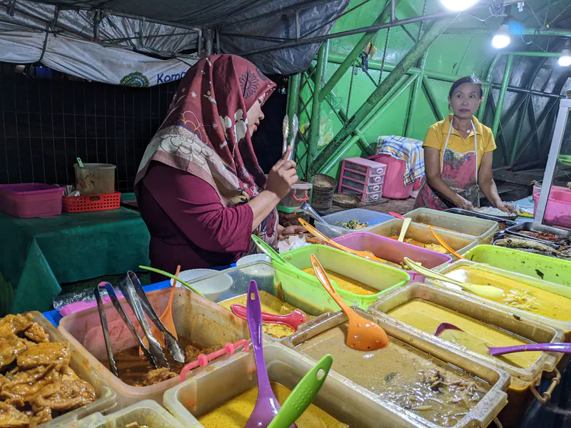 Two women at a food stall with trays of curry and side dishes.