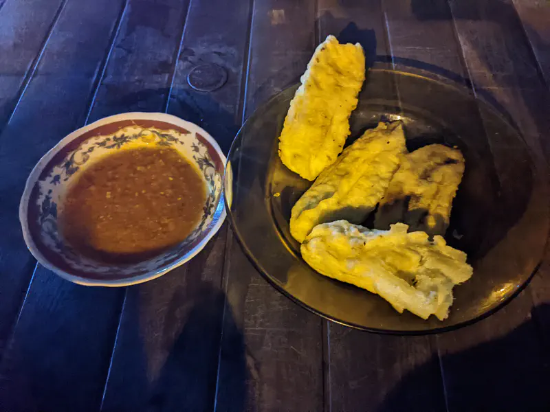 A plate of fried banana fritters served with a bowl of spicy dipping sauce on a dark wooden table.