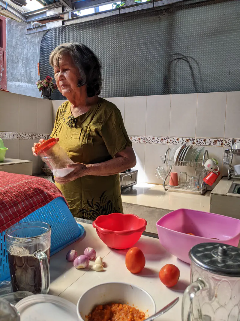 Elderly woman in a kitchen holding a container of salt near vegetables on a table.
