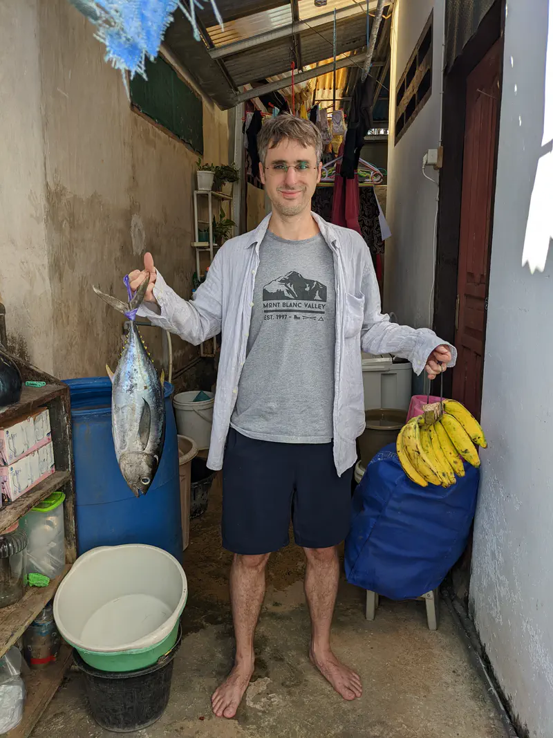 Man standing barefoot in a narrow outdoor corridor, smiling while holding a large fish in one hand and a bunch of bananas in the other.