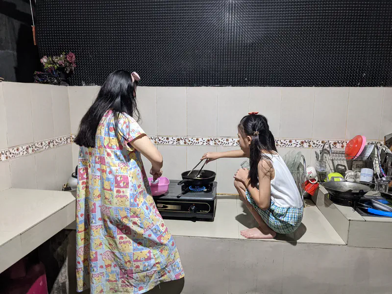 Two girls cooking together in a simple tiled kitchen, one standing and stirring while the other squats beside the stove using a spatula, with dishes stacked on a drying rack nearby.