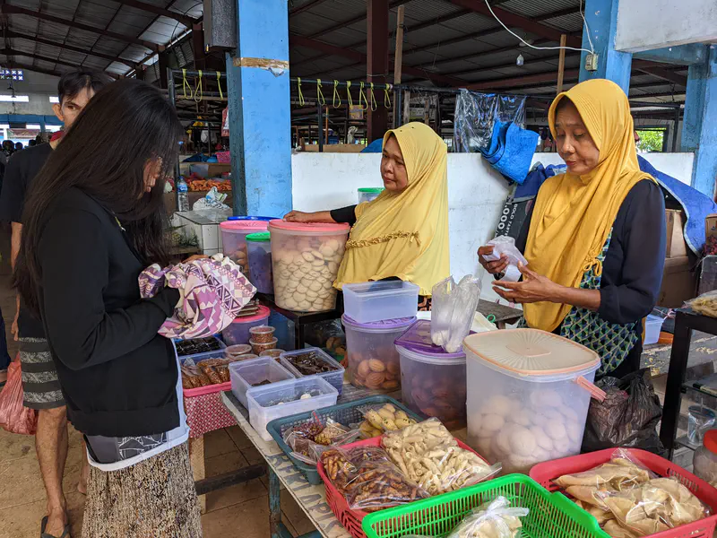 A woman buying snacks from two vendors in yellow hijabs at a traditional market stall filled with jars and baskets of local treats.