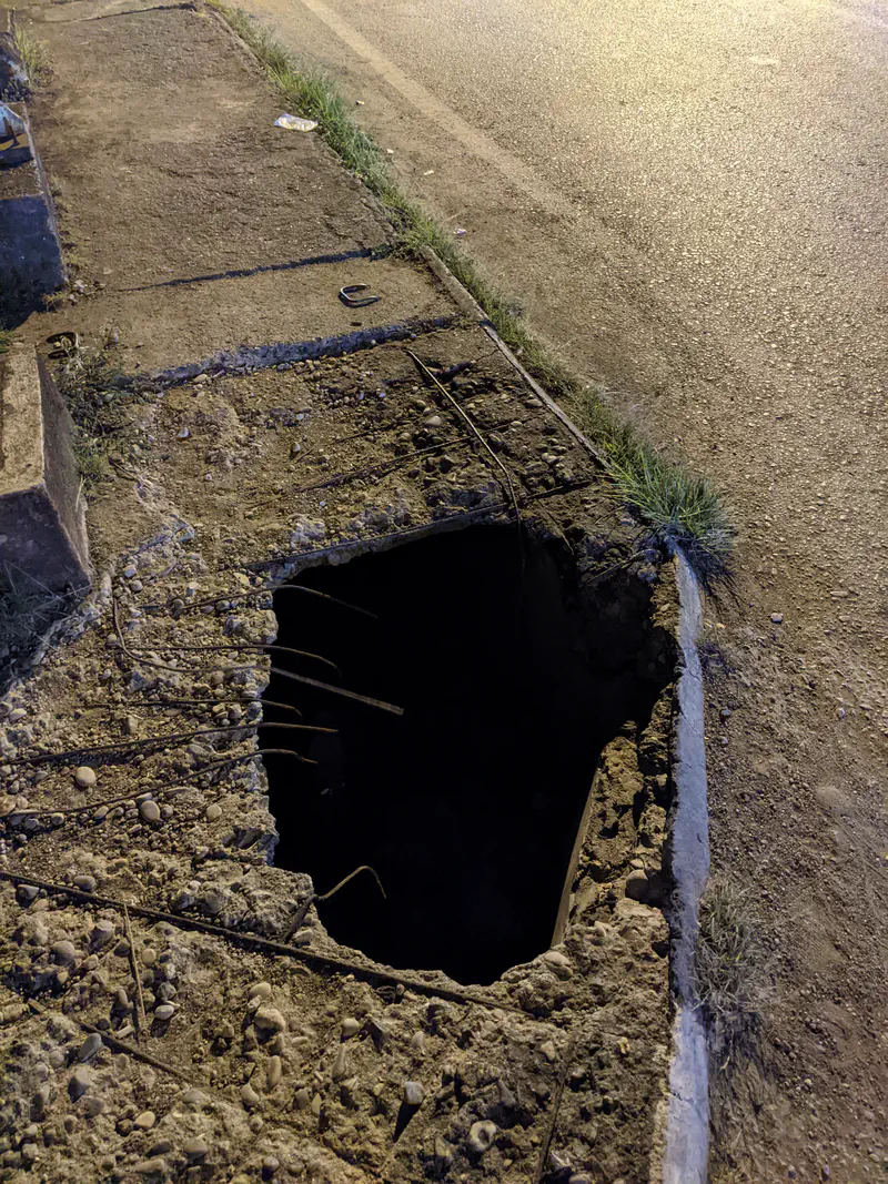 Large hole in a damaged sidewalk near the road at night.