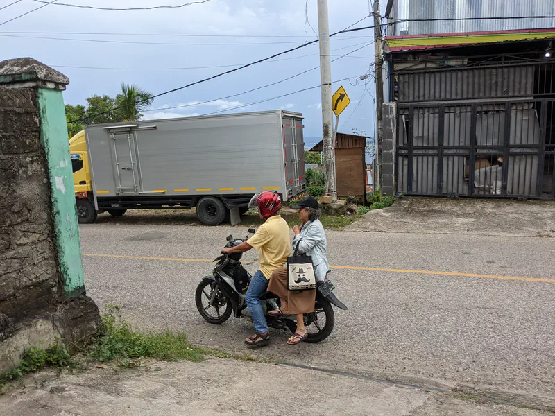 Two people riding a motorbike on a street with a truck passing by.