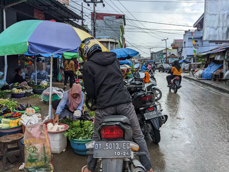 Busy street market with vegetable vendors under umbrellas and motorcycles parked nearby.