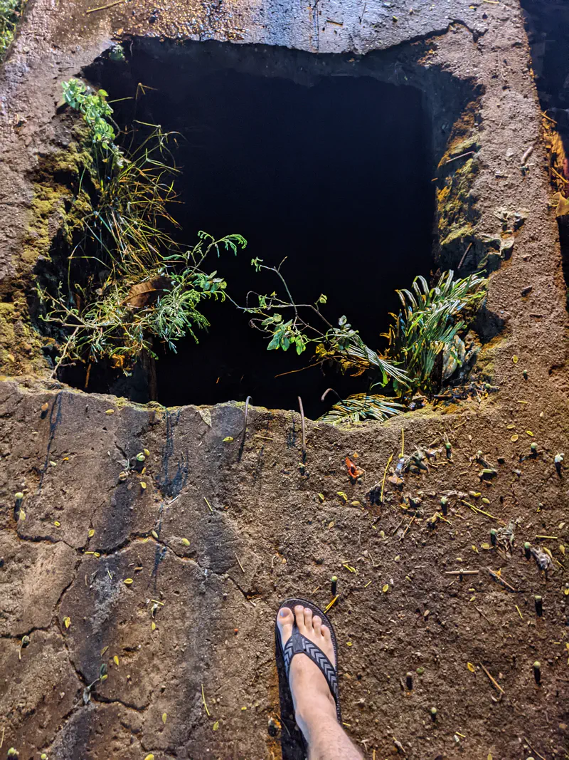 Open hole in a sidewalk with a foot in sandals standing nearby.