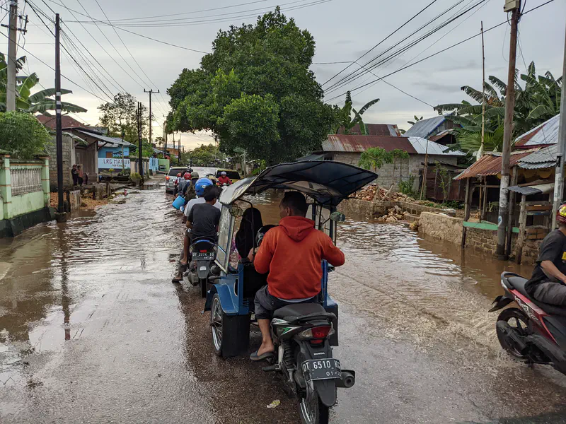 Motorcycles and a rickshaw driving through a flooded street in a village.