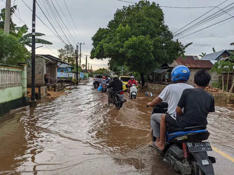 People riding motorcycles through floodwater on a village street.