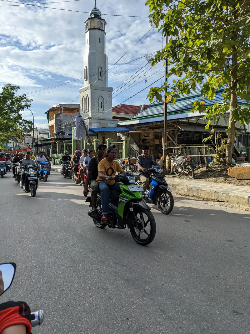 Group of people riding motorcycles past a mosque with a tall white minaret.