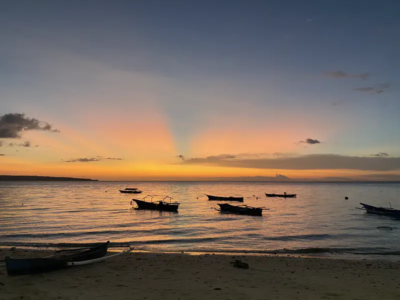 Colorful sunset over the ocean with small boats silhouetted on the calm water and sandy beach in the foreground.