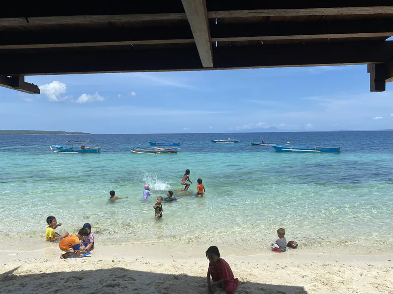 Children playing in the shallow turquoise water and on the sandy beach, with fishing boats floating in the background.