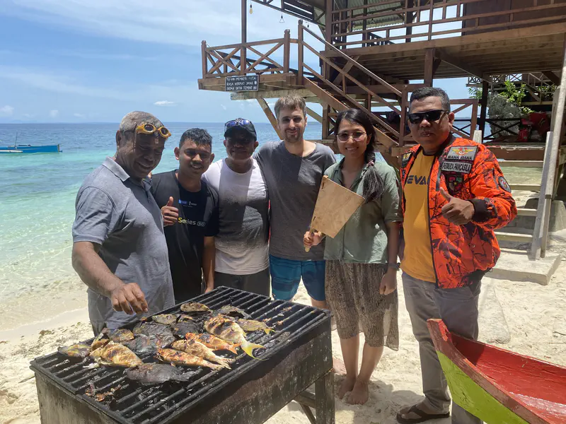 Group of people standing by a barbecue with grilled fish at the beach.