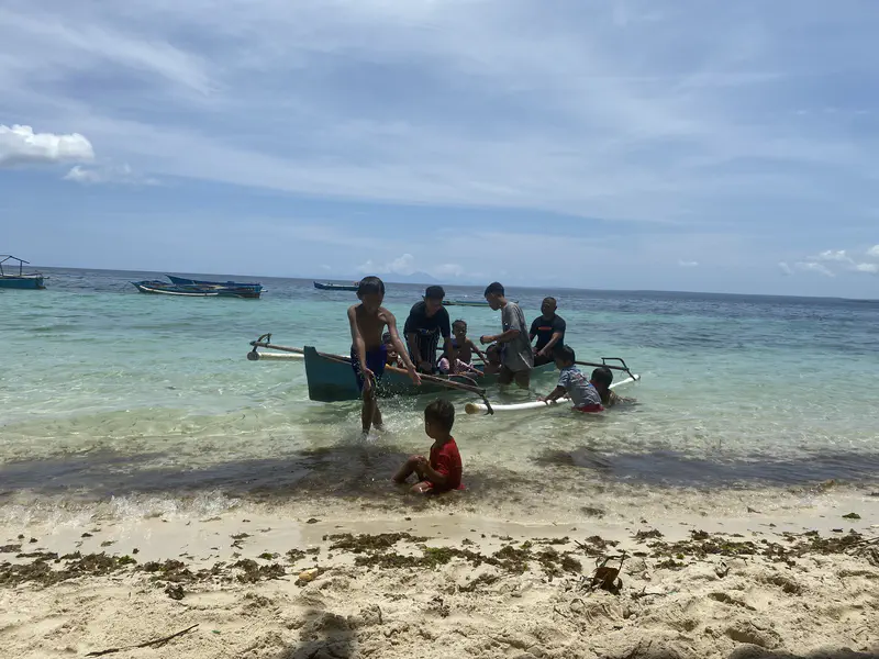 Children playing with a small boat in shallow turquoise water, while one child sits on the sandy beach.