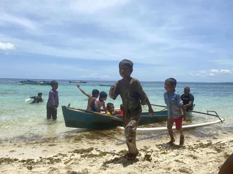 Children playing with a small boat on the beach, some in the water and others on the sand, under a sunny sky.