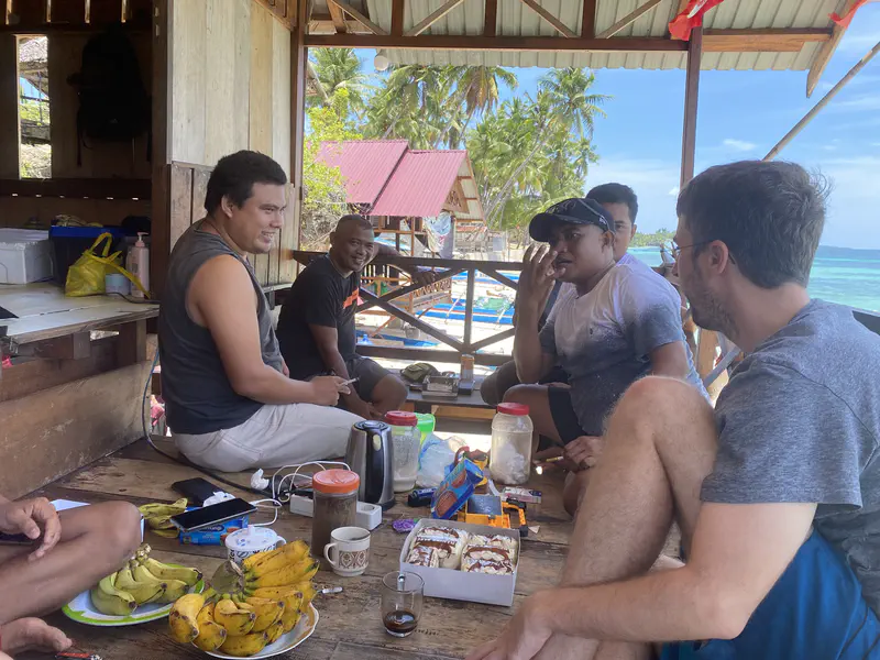 Group of people sitting on a wooden porch sharing food and drinks by the beach.