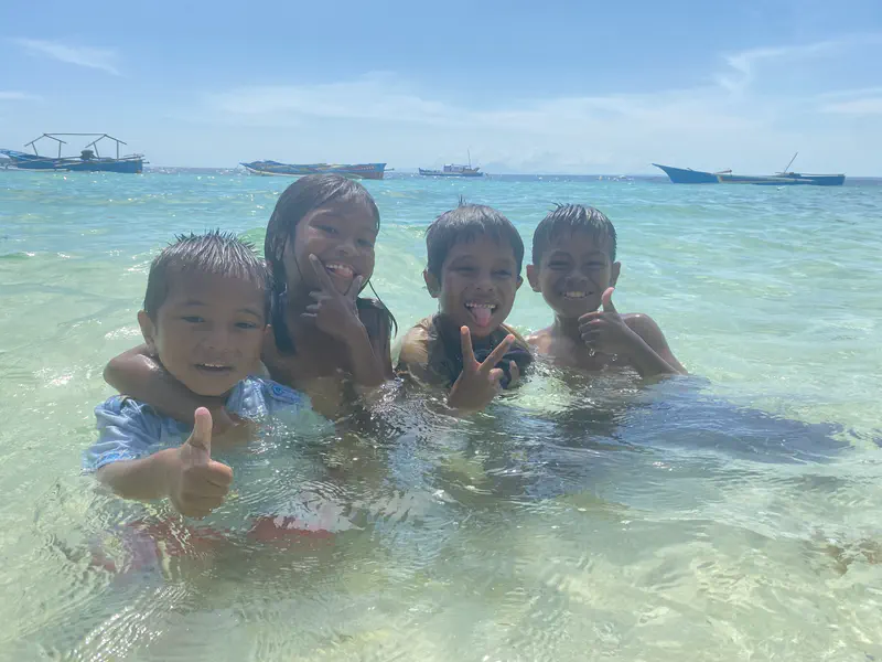 Four children smiling and posing with thumbs up and peace signs while playing in clear shallow seawater, with boats in the background.