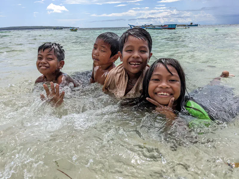 Four smiling children playing in shallow seawater with boats visible in the distance.