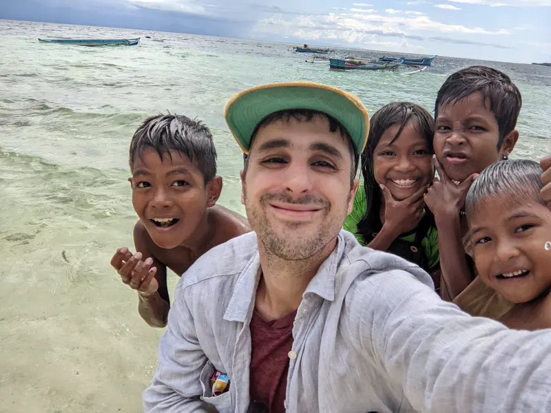 Smiling man taking a selfie with children on a beach, with boats floating on the sea in the background.