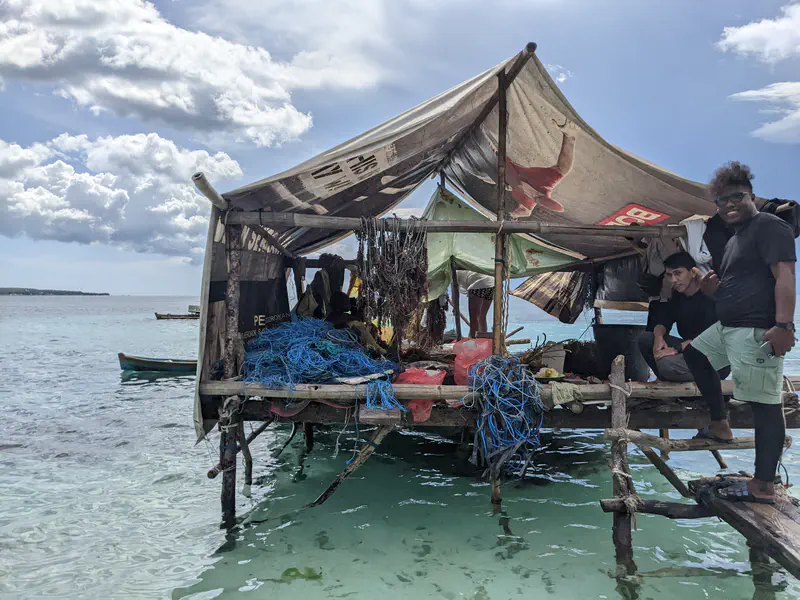 People working on a stilt hut above the water, processing seaweed with ropes and nets scattered around, while others sit and smile nearby.