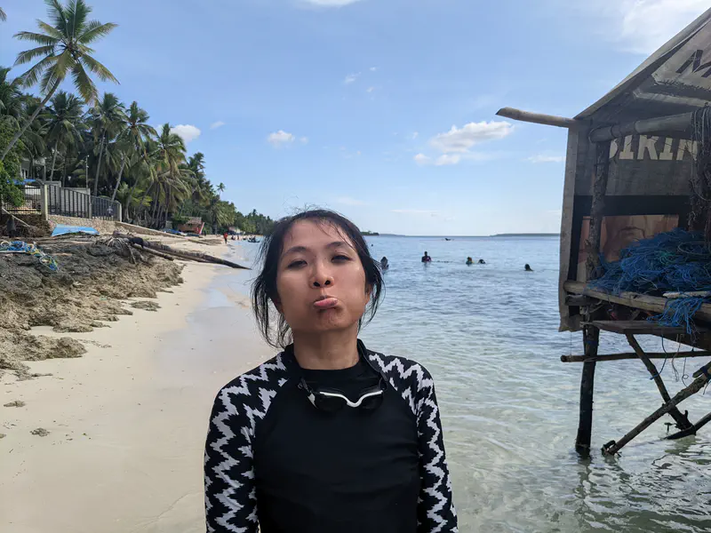A woman in a black and white rash guard stands on a tropical beach making a playful pout, with palm trees and a stilt hut over the water in the background.