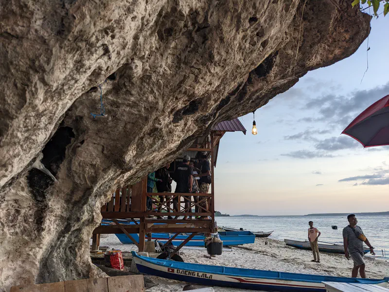 Beachside scene at sunset with people near wooden huts built under a rocky cliff, fishing boats on the sand, and the sea in the background.