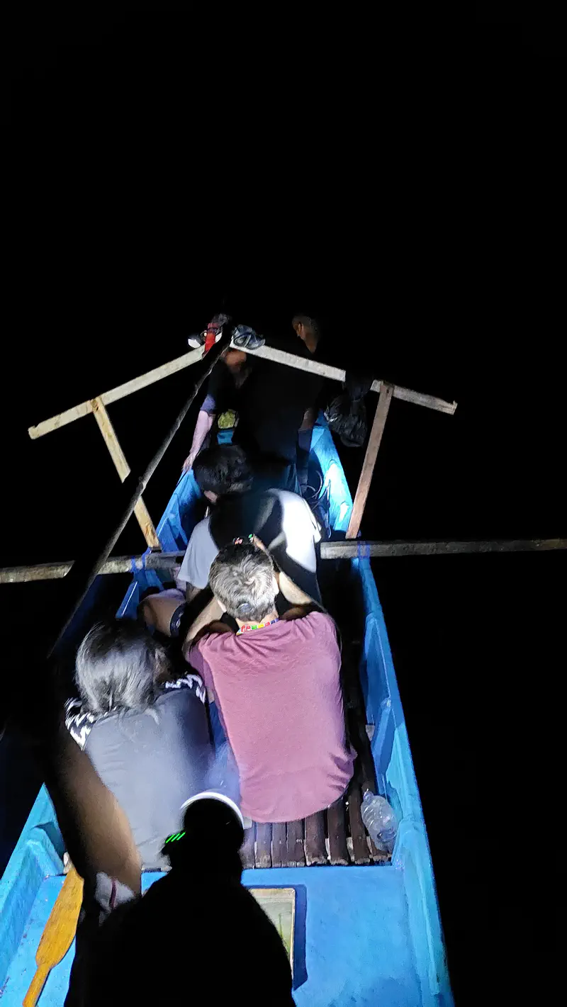 People sitting close together in a small blue outrigger boat at night, illuminated by headlamps and flashlights against the dark sea.