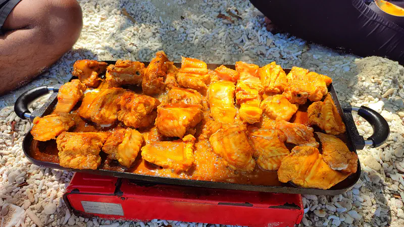 Marinated fish pieces grilling on a portable stove over coral sand.