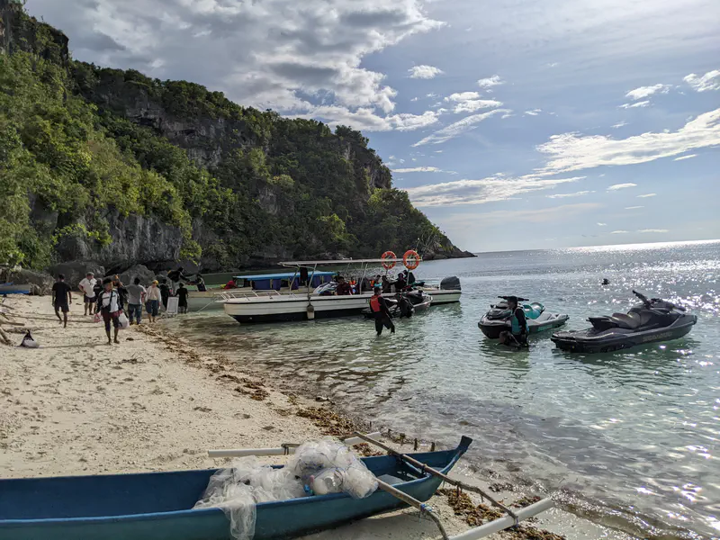 People arriving at a beach with speedboats and jet skis anchored in clear water, surrounded by rocky cliffs and lush greenery.