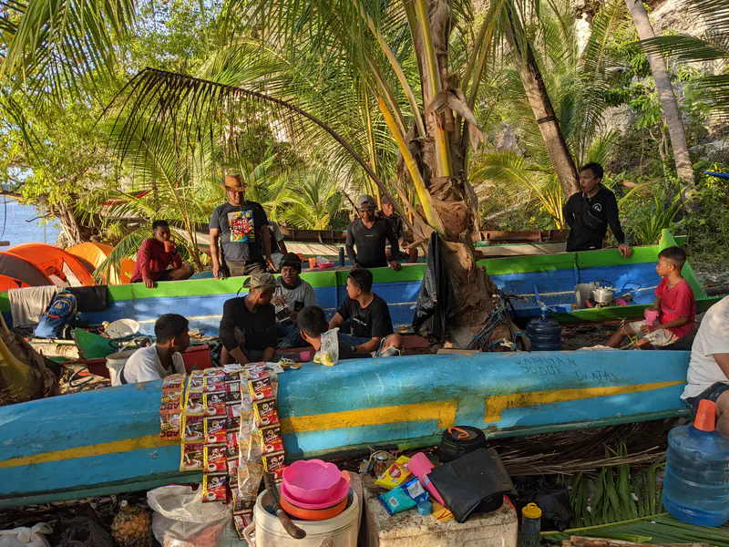 Group of people gathered in a shaded area by the beach, sitting and standing around colorful wooden boats repurposed as seating and tables, with food and supplies spread out.