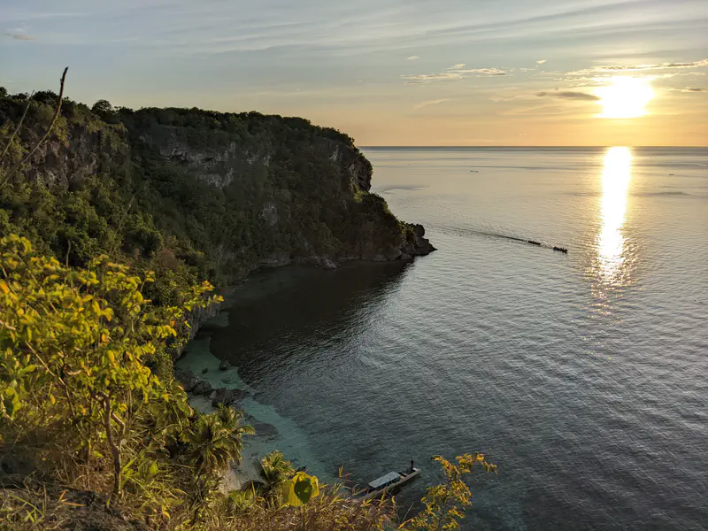 Person standing on a rocky path framed by cliffs, looking out over a turquoise bay surrounded by lush greenery and cliffs.