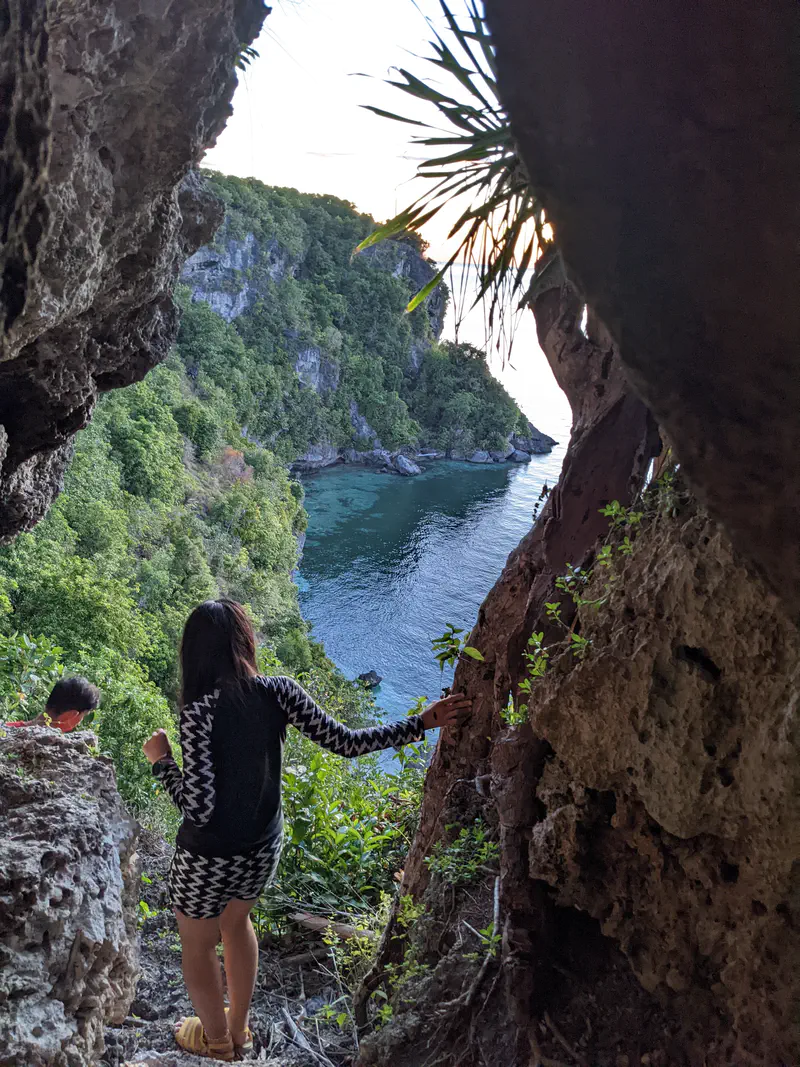 Person standing on a rocky path framed by cliffs, looking out over a turquoise bay surrounded by lush greenery and cliffs.