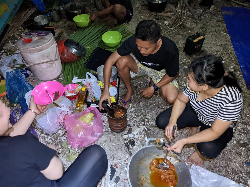 Group of people sitting on the ground outdoors at night, preparing food together with bowls, cooking pots, and ingredients spread around them.