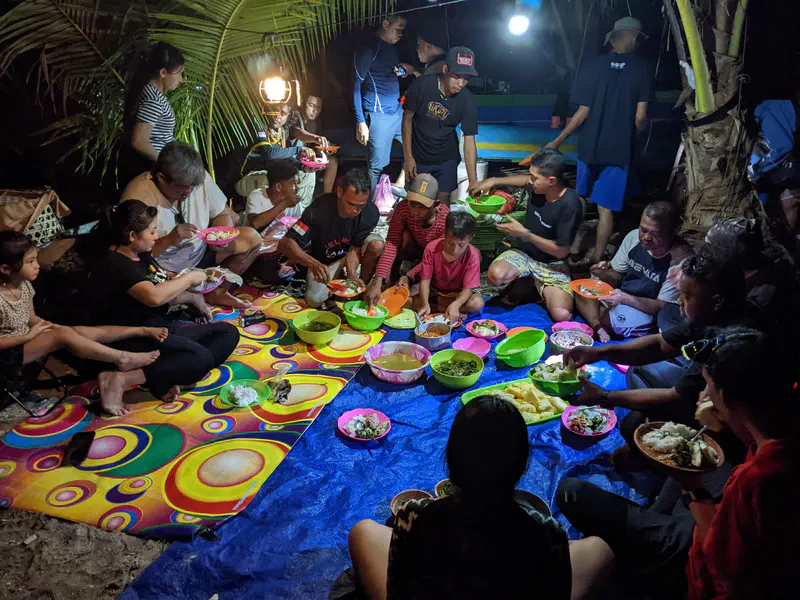 Outdoor communal dinner at night with bowls of rice, vegetables, soup, pineapple, and other dishes placed on a tarp, as people sit in a circle sharing the meal together.