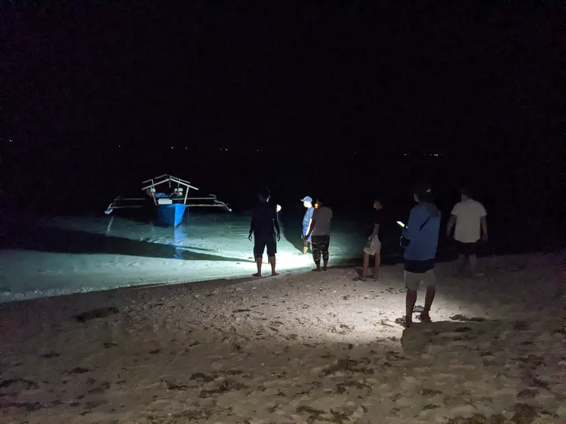 Group of people standing on a sandy beach at night with flashlights, looking toward a small blue outrigger boat anchored in shallow water.