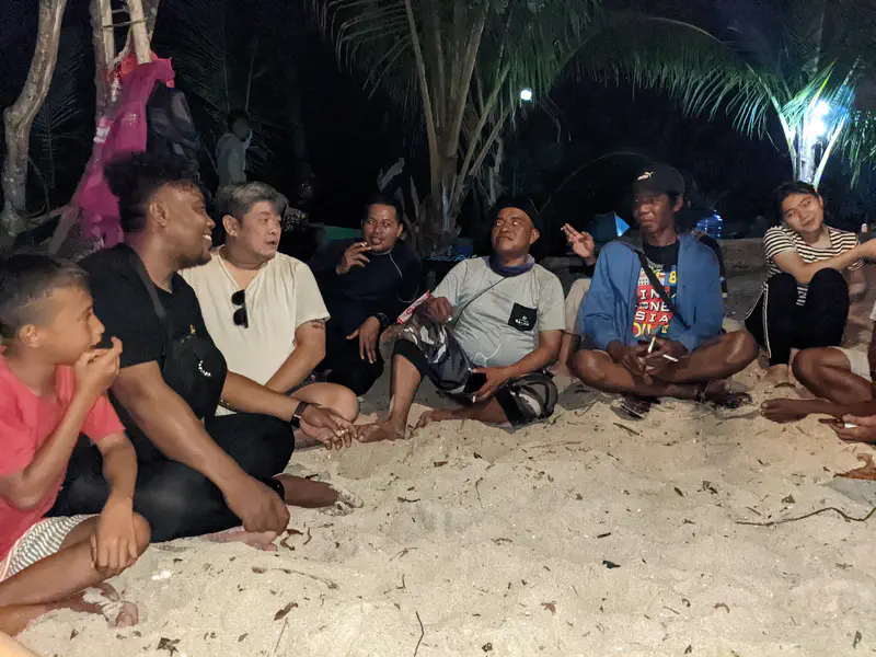 Group of people sitting in a circle on sandy ground at night, casually talking and laughing under the light of nearby lamps and palm trees.