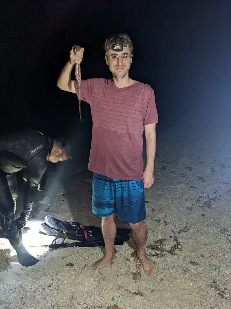 Man wearing a headlamp standing on a beach at night, smiling and holding up a small octopus, with another person in a wetsuit working under flashlight nearby.