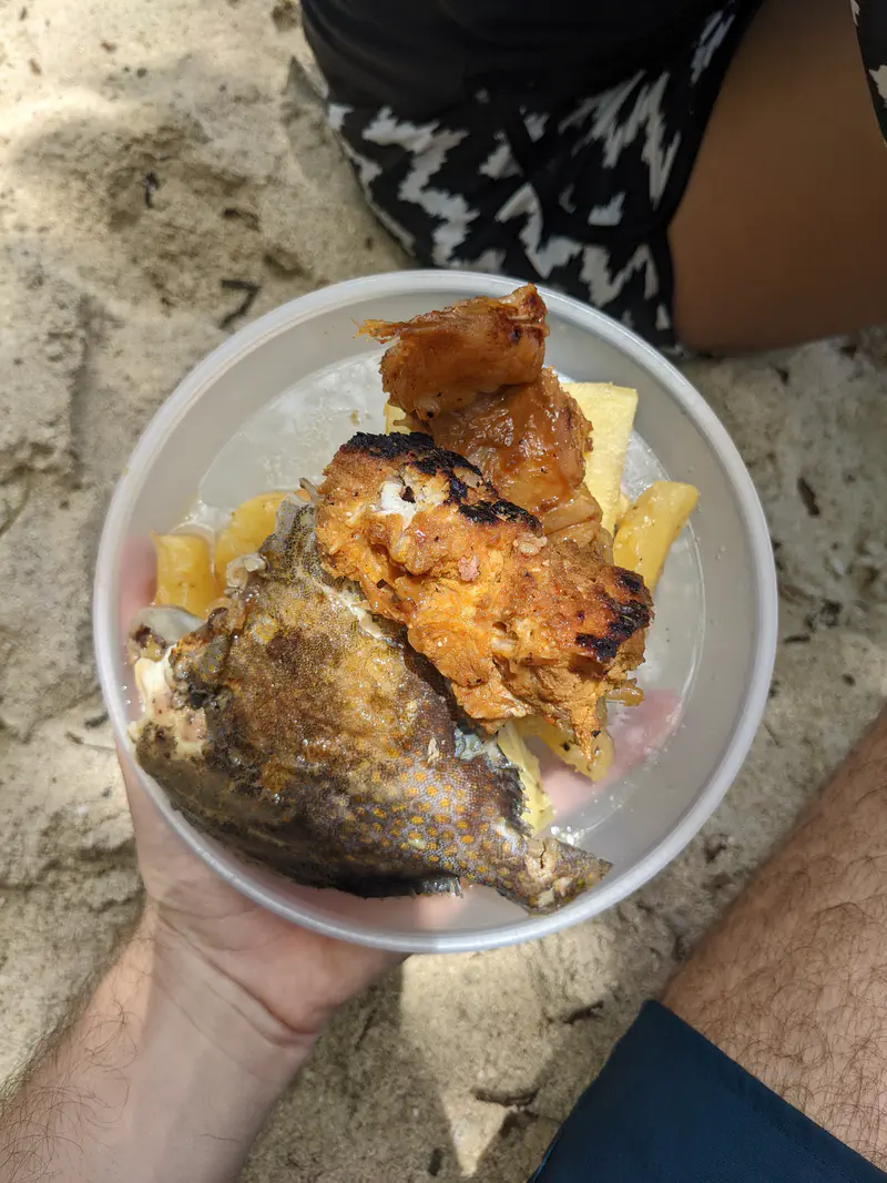 Bowl with grilled fish, fried fish pieces, and boiled cassava on sand.