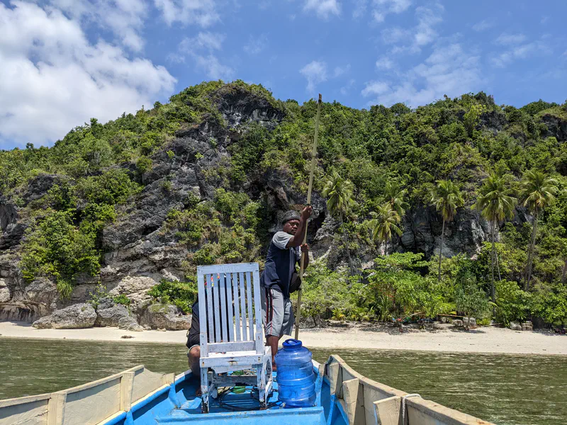 Man standing on a blue boat holding a wooden pole near a tropical shoreline with cliffs and palm trees.