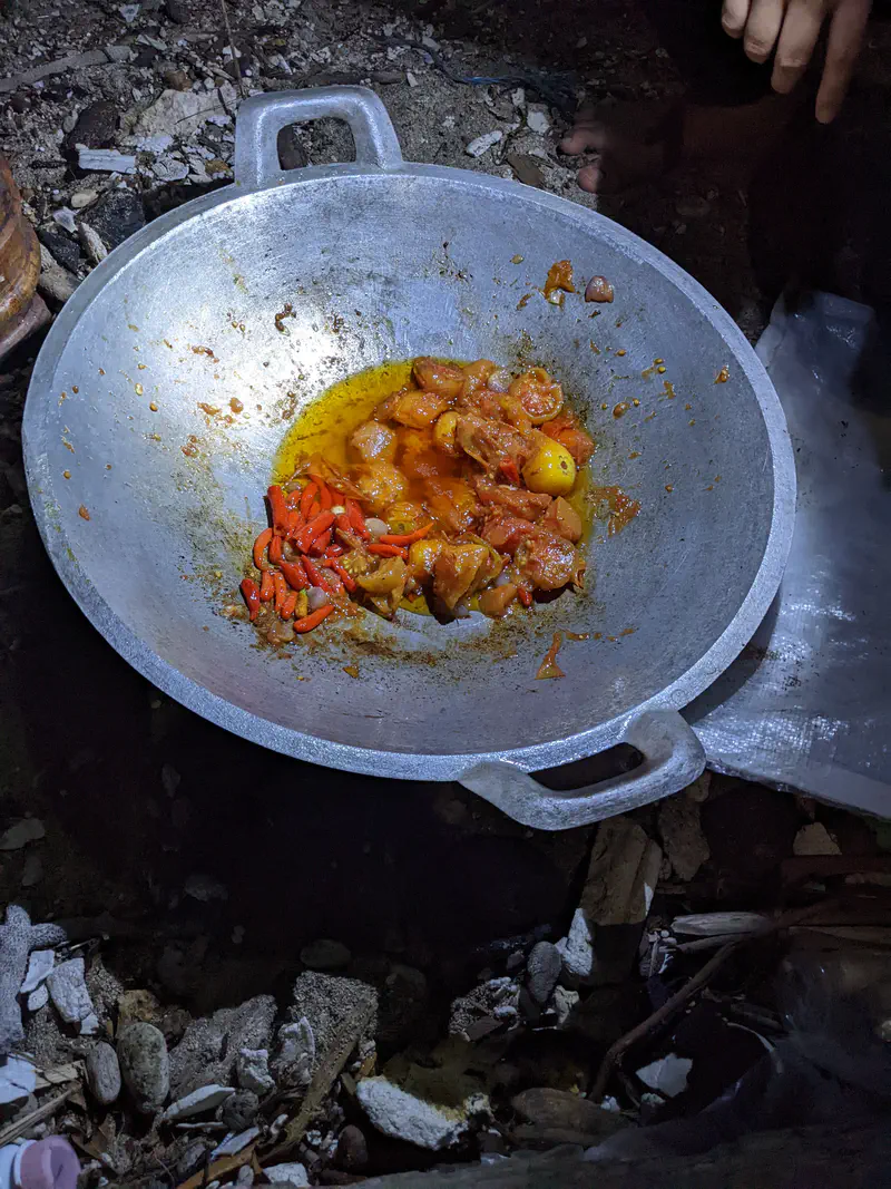 Large metal pan on the ground containing a spicy dish with red chili peppers, potatoes, and meat in an oily sauce, prepared outdoors at night.