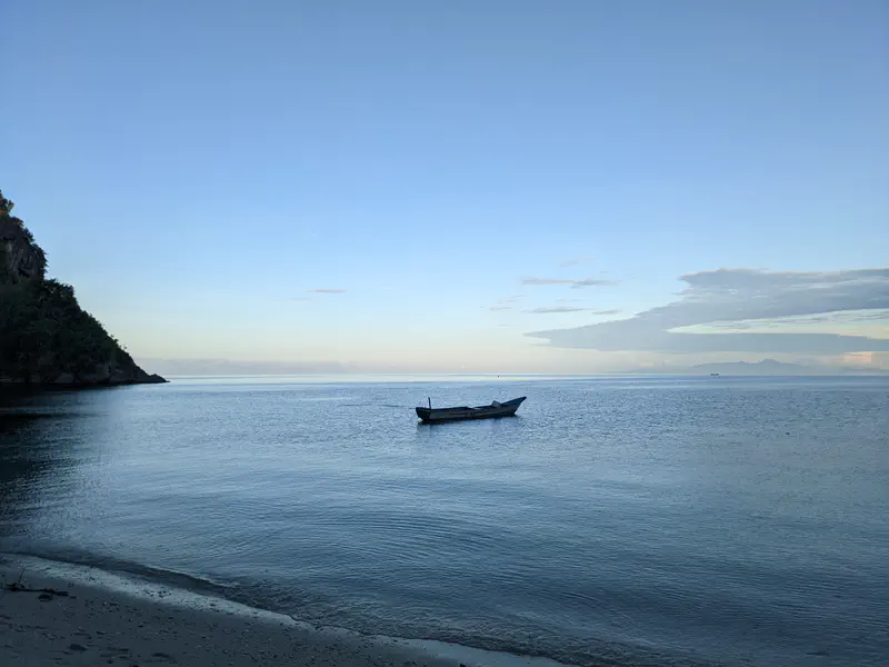 Single wooden boat floating on calm sea at dawn with soft blue sky and distant mountains on the horizon.