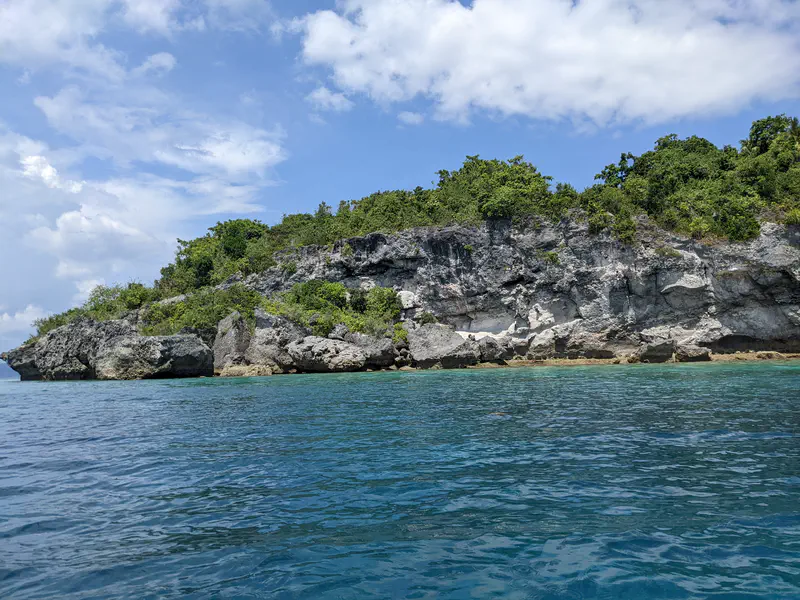 Rocky coastline with green vegetation above clear turquoise water under a partly cloudy sky.